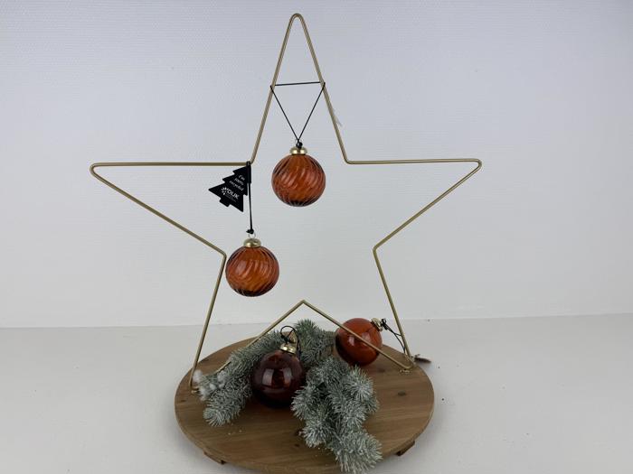 En stjerne i guldtråd står på en træbase, dekoreret med orange glaspynt og fyrretræsgrene.