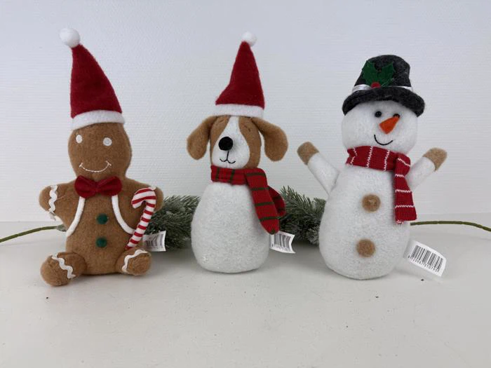 Tre plysdyr: en honningkagemand med sukkerstang, en hund og en snemand, alle iført festlige hatte og tørklæder, sidder foran en fyrretræsgren på en hvid baggrund.