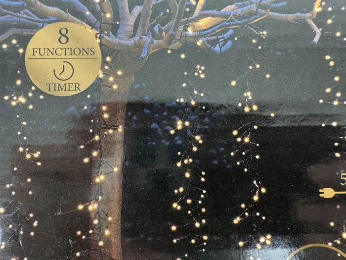 Et træ pyntet med glødende lyskæder om natten, med en guldlabel med 8 funktioner og timer i øverste venstre hjørne.