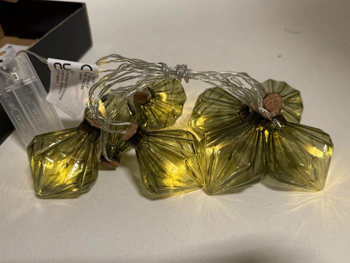 En række gulgrønne glasflaskeformede lyskæder med korkpropper, der er tændt og hviler på en hvid overflade.