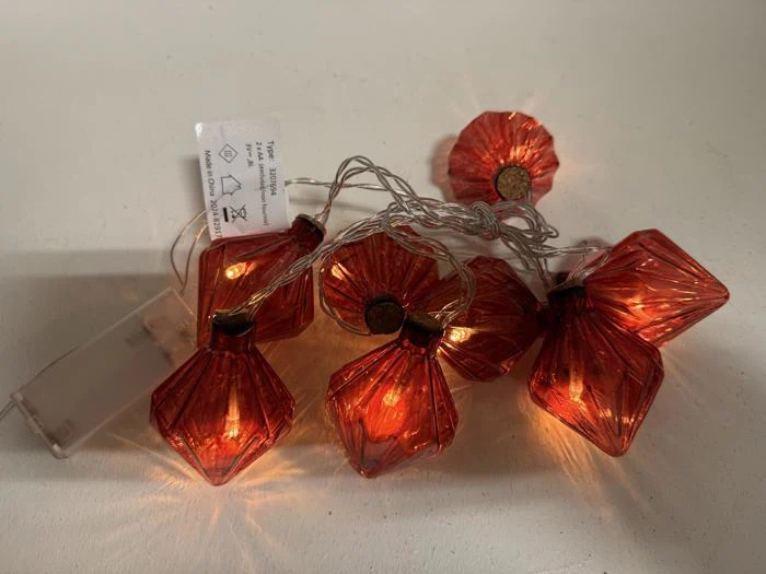 En række tændte orange diamantformede lyskæder med en klar batteripakke, der hviler på en hvid overflade.