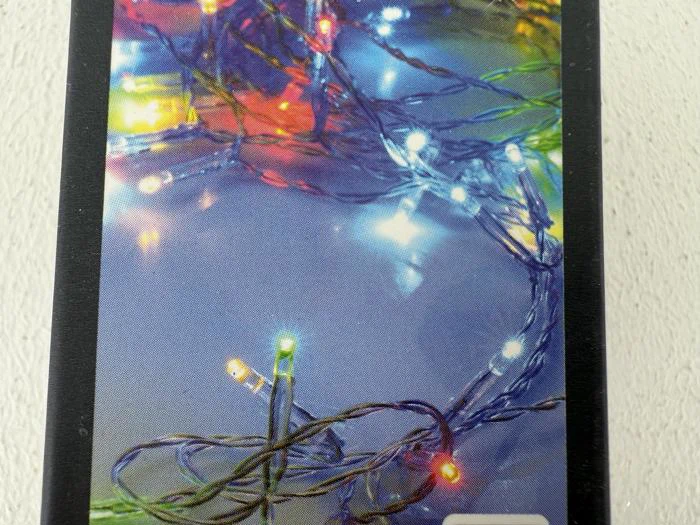 Farverige lyskæder med røde, gule, grønne og blå pærer er viklet sammen og lyser op mod en lys baggrund.