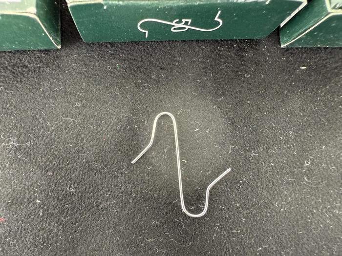 En enkelt sølvfarvet metalkrog ligger på en sort struktureret overflade foran grønne kasser med hvide motiver.