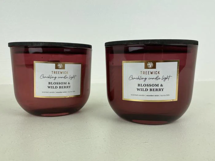 To røde glaslys med mørke låg, mærket Trewick Crackling Candle Light, Blossom & Wild Berry, på en hvid flade mod en lys baggrund.