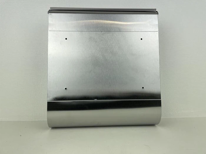 En skinnende, rektangulær metaldispenser med en buet bund, monteret mod en lys struktureret væg. Fire små huller er synlige på den flade forside.