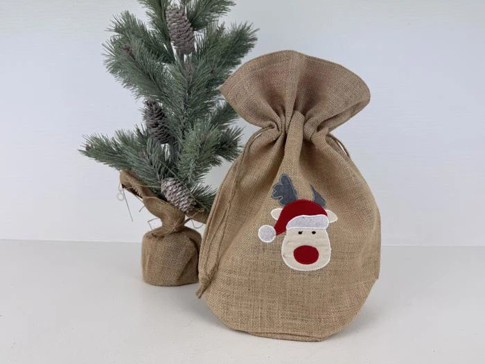 En lille gavepose i jute med et rensdyr med nissehue står ved siden af et lille kunstigt juletræ pakket ind i jute.