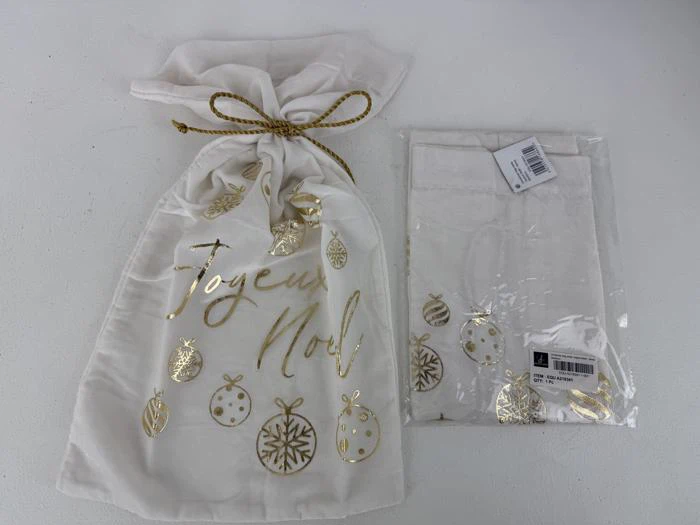 En hvid gavepose med snor med Joyeux Noël og guldornamenter, ved siden af en matchende foldet pose i klar emballage.