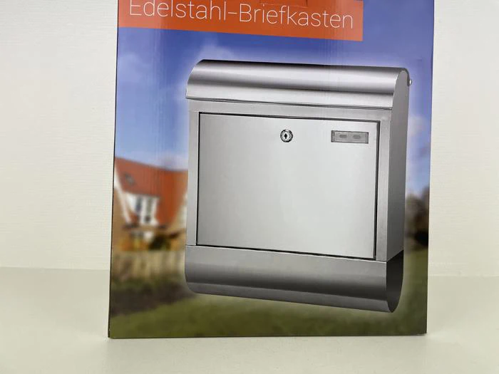 En postkasse i rustfrit stål med lås og navneskilt på en sløret baggrund af et hus og en have. Teksten ovenover lyder Edelstahl-Briefkasten.