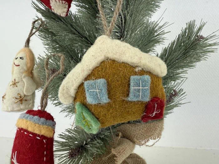 Et hus i filt med blå vinduer og hvidt tag hænger på en fyrretræsgren ved siden af andre dekorationer i filt, bl.a. en rød vante og en smilende engel.