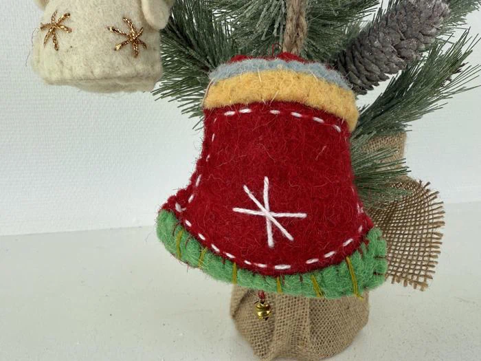 En håndlavet rød filtklokke med hvide syninger og grønne og gule detaljer hænger på en fyrregren med sækkelærred og kogler.