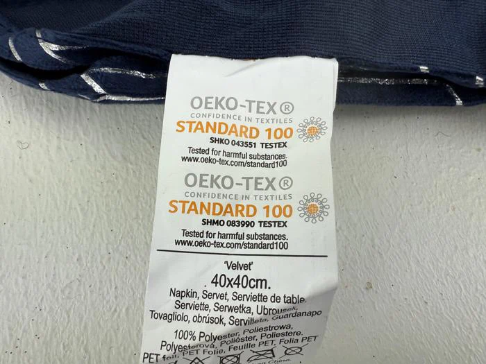 En stofetiket viser OEKO-TEX STANDARD 100-certificeringsoplysninger, vareinformation og materialesammensætning for en 40x40 cm fløjlsserviet på flere sprog.