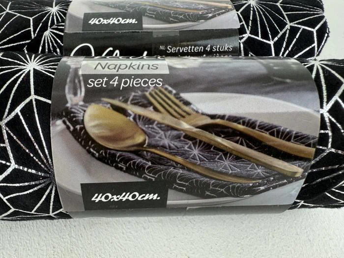 En pakke med fire sorte servietter med et hvidt geometrisk mønster, mærket Serviettsæt 4 stk. 40x40 cm, med et billede af servietter, en gaffel, en kniv og en ske.