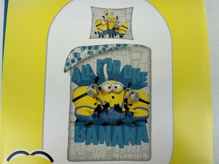 En dobbeltseng med sengetøj med Minions-tema, hvor der står ALL FOR ONE BANANA med tre Minions, der holder bananer, på en gul baggrund.