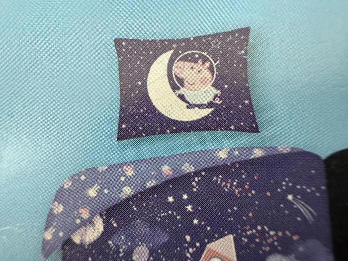 En pude med et tegneseriebarn, der sidder på en halvmåne omgivet af stjerner på en mørkeblå baggrund. Matchende sengetøj med stjerner er delvist synligt nedenfor.