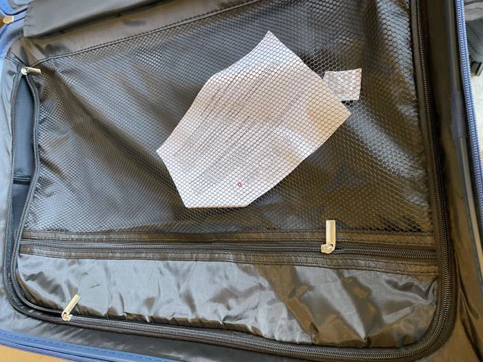 En hvid ansigtsmaske med gittermønster ligger i en netlomme i en åben sort kuffert.