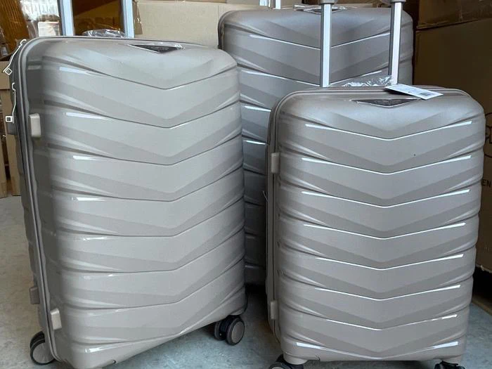 Tre beige hard-shell kufferter med diagonale mønstre står oprejst, to med forlængede håndtag og alle med hjul, placeret indendørs i nærheden af papkasser.