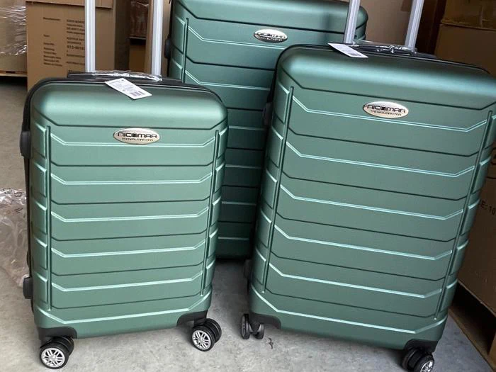 Tre grønne hard-shell kufferter i forskellige størrelser med hjul, der står oprejst sammen på et gulv. Hver kuffert har en sølvfarvet logoplade på forsiden og et hvidt bagagemærke på håndtaget.