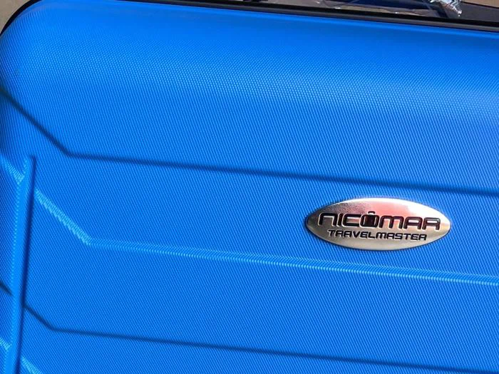 Nærbillede af en blå kuffert med en metallisk, oval etiket med teksten NICOMAR TRAVELMASTER.