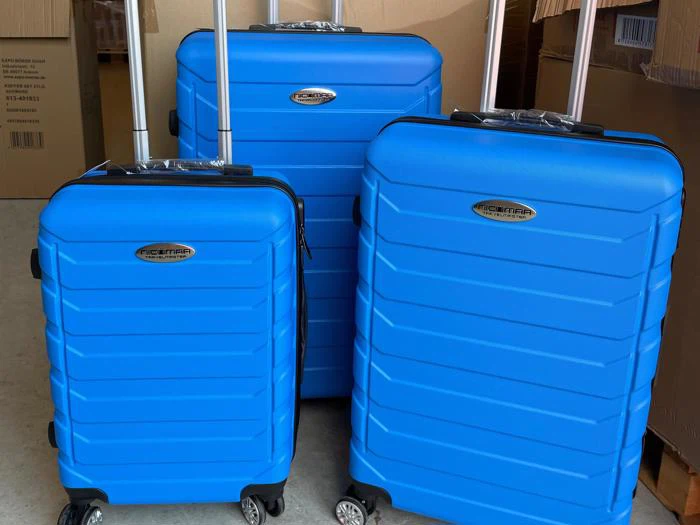 Tre blå hardshell-kufferter i forskellige størrelser med teleskophåndtag og hjul står side om side, omgivet af papkasser.
