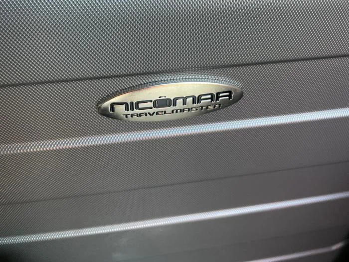 Nærbillede af en struktureret grå overflade med et ovalt metallogo, hvor der står NICOMAR TRAVELMASTER.