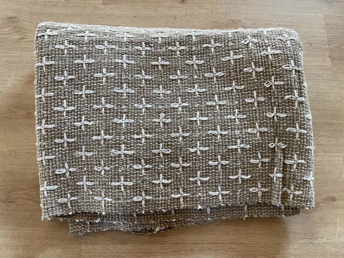 Et foldet brunt tekstureret tæppe med et mønster af små hvide korssting, placeret på en træoverflade.
