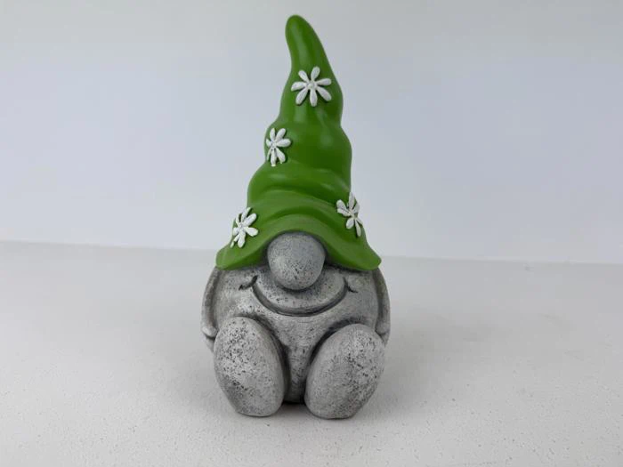 En lille grå nissefigur med et stort smil og en høj grøn hat dekoreret med hvide blomster, der sidder på en lys overflade.
