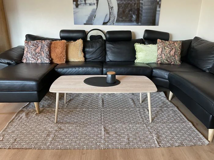 En sort lædersofa med forskellige puder står bag et sofabord i lyst træ på et mønstret tæppe i en stue. Over sofaen hænger en sort-hvid vægkunst.