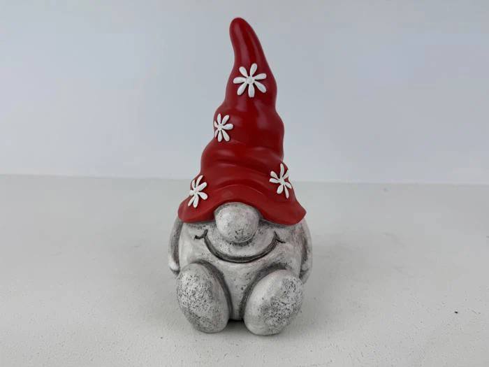 En lille, smilende nissefigur med en høj rød hat dekoreret med hvide blomster, der sidder med knæene oppe på en glat overflade mod en lys baggrund.