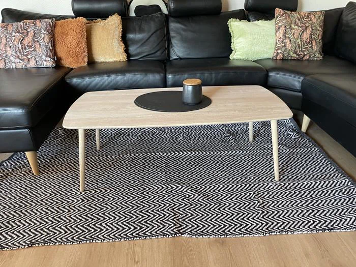 Et sofabord i lyst træ med en sort bakke og kop står på et sort-hvidt zigzag-tæppe foran en sort sektionssofa med mønstrede og strukturerede puder.