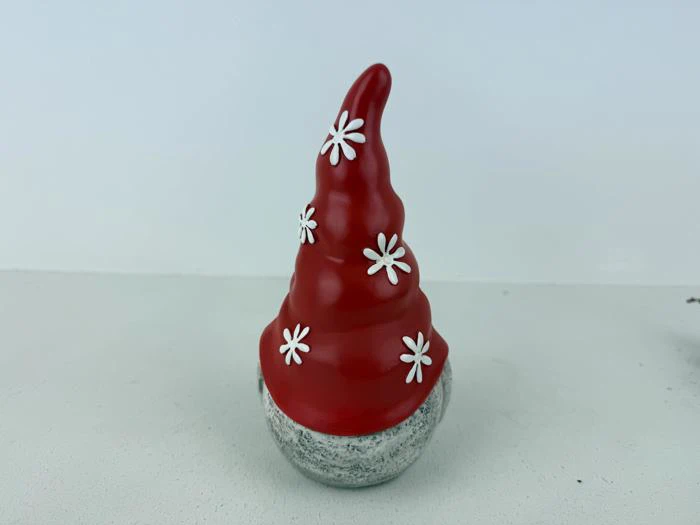 En lille figur med en høj, rød, kegleformet hat dekoreret med hvide blomster, der sidder på en rund, grå base mod en ensfarvet baggrund.