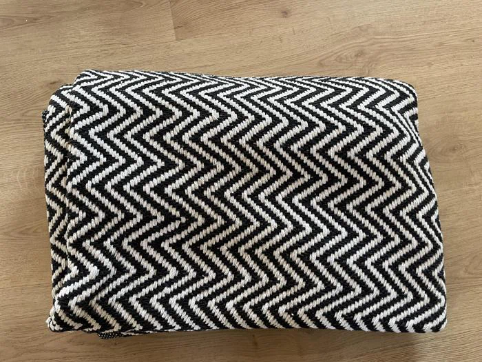 Et foldet sort-hvidt tæppe med et fedt zigzag-chevrondesign ligger på et lyst trægulv.