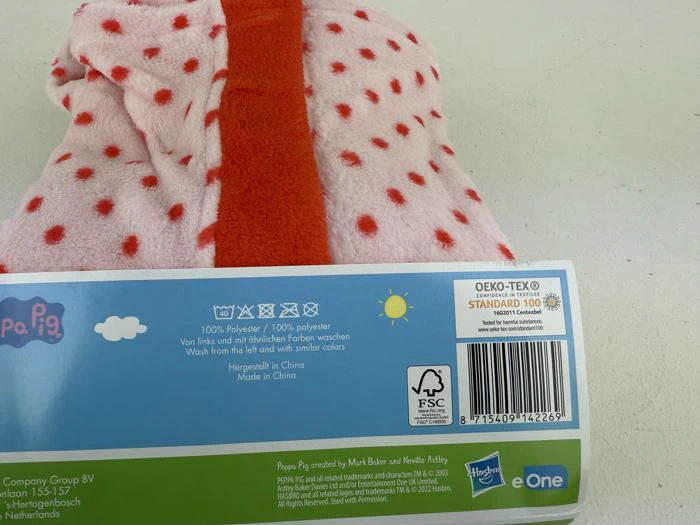 En pink og rød prikket fleecetrøje med en produktetiket, der viser plejeanvisninger, certificeringer og en stregkode på en hvid overflade.