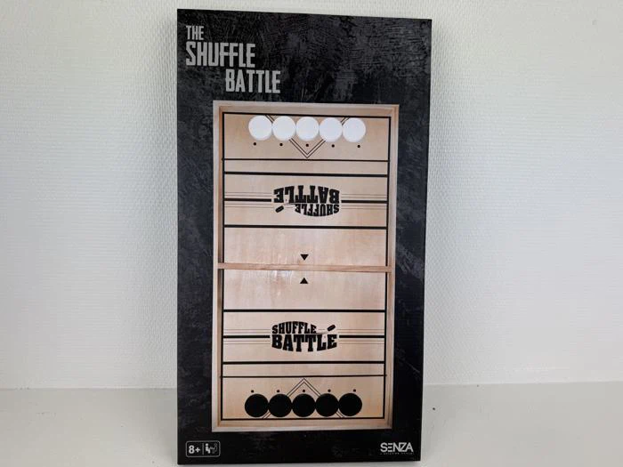 En rektangulær æske med spillet The Shuffle Battle, der viser en træplade med pucker og spilmarkeringer på forsiden. Egnet til børn fra 8 år og opefter.