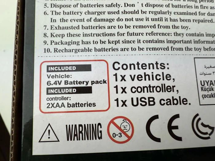 Nærbillede af produktemballagen, der viser de medfølgende elementer: 1 køretøj, 6,4 V batteripakke, 2 AA-batterier, 1 controller, 1 USB-kabel, med advarselsskilte for sikkerhed og aldersbegrænsning (0