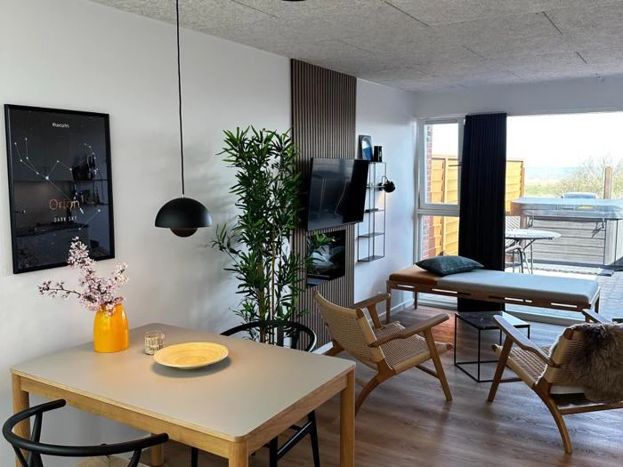 Et moderne, hyggeligt opholds- og spiseområde med træmøbler, et lille spisebord, lænestole, et væghængt tv, store vinduer og planter med udsigt til en gårdhave udenfor.