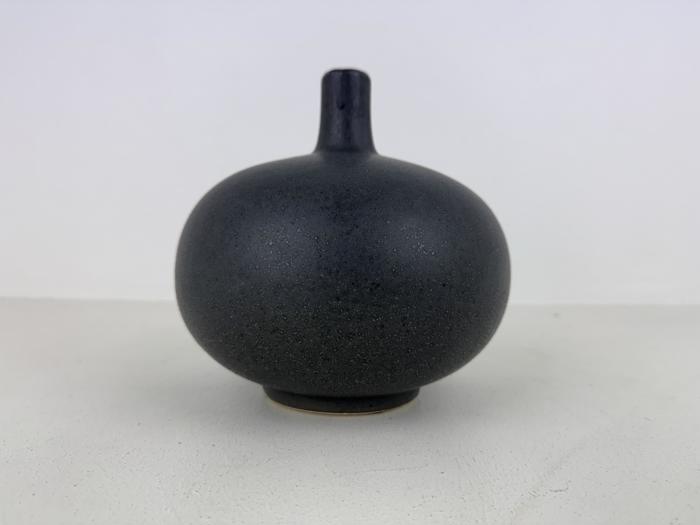 En lille, rund, mørkegrå keramikvase med smal hals, placeret på en lys overflade mod en almindelig hvid baggrund.