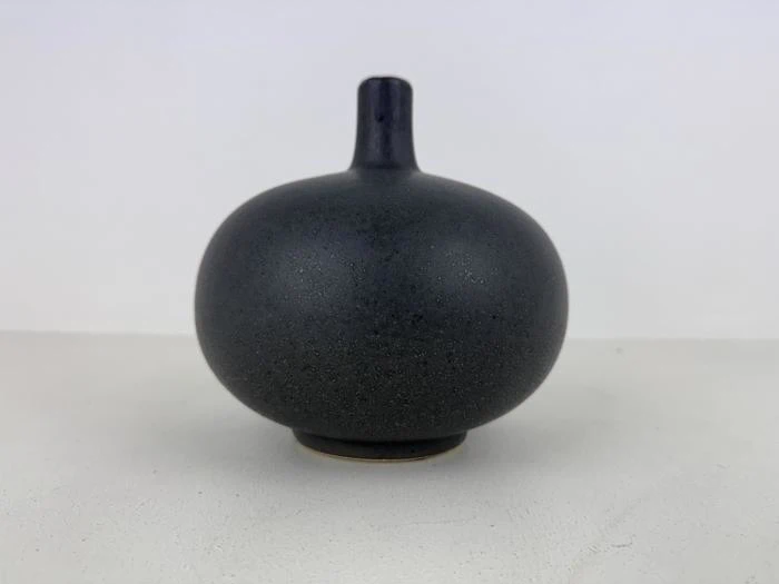 En lille, rund, mørkegrå keramikvase med smal hals, placeret på en lys overflade mod en almindelig hvid baggrund.