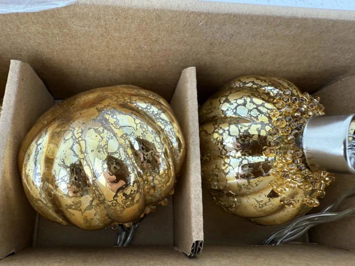 To græskarformede ornamenter af guld og glas med prikkede mønstre er adskilt af papskillevægge inde i en æske. Det ene ornament har dekorative perler nær stilken.