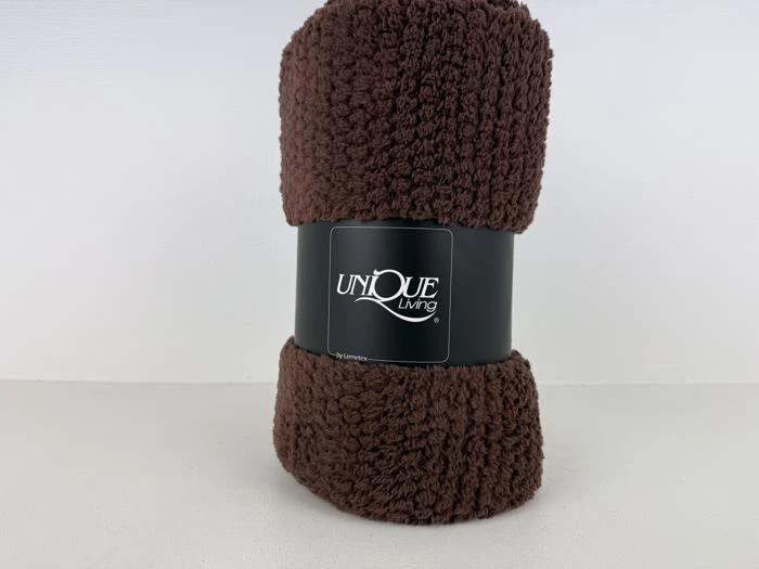 Et sammenrullet, mørkebrunt, fluffy tæppe med en sort etiket, hvor der står Unique Living, på en almindelig hvid baggrund.