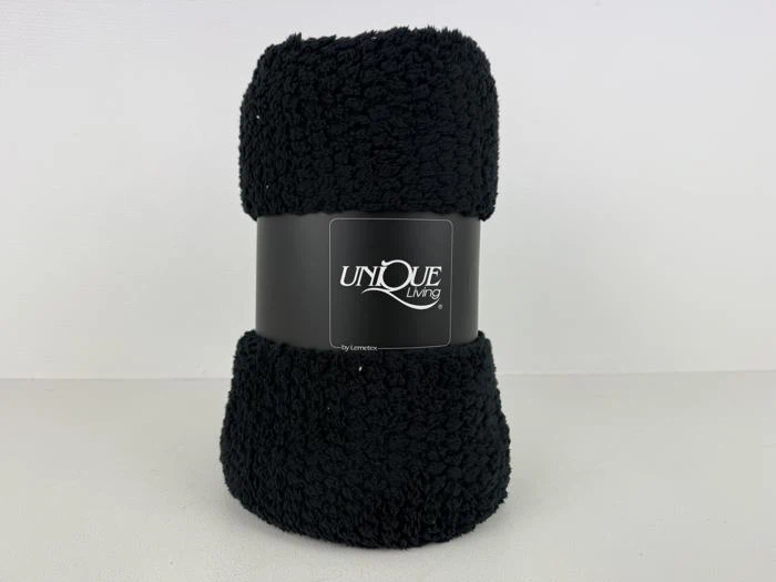 Et sammenrullet sort fleecetæppe med en sort etiket med teksten UNIQUE Living, placeret på en almindelig hvid overflade.