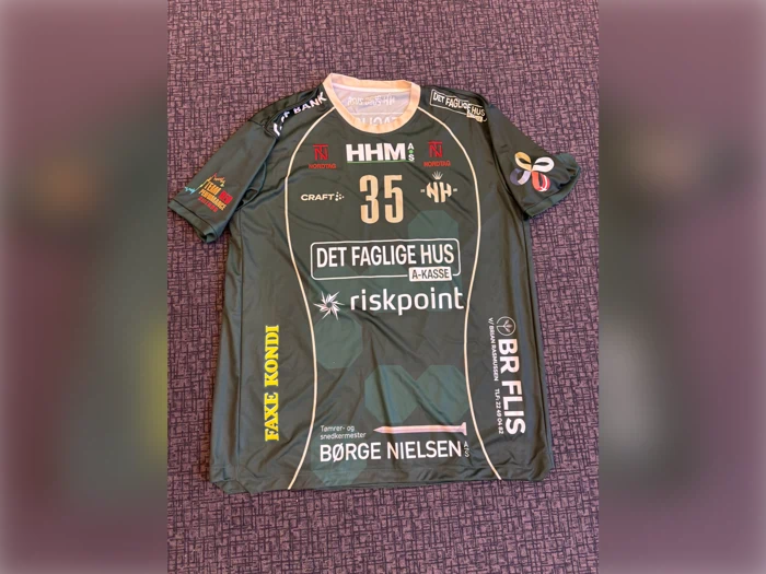 Grøn sportstrøje med nummer 35, flere sponsorlogoer og teksten »DET FAGLIGE HUS«. Trøjen er udstillet på en lilla, struktureret overflade.