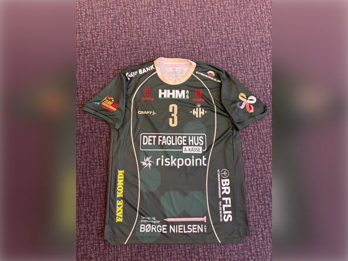 En grøn sportstrøje med nummer 3, forskellige sponsorlogoer og hvid kant ligger fladt på en lilla mønstret overflade.