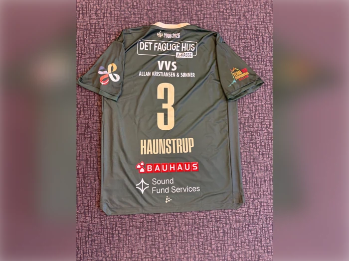 En grøn sportstrøje med nummer 3 og navnet Haunstrup på ryggen, med flere sponsorlogoer på en mønstret lilla baggrund.