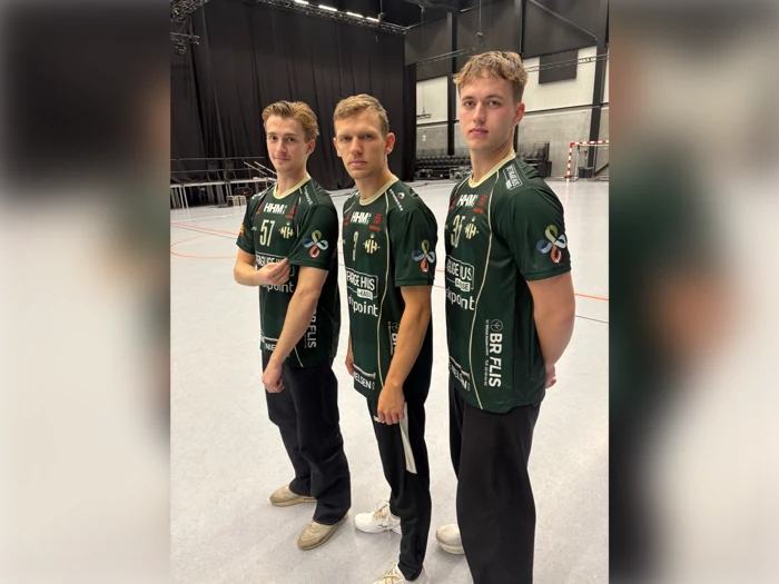 Tre unge mænd i ens grønne sportstrøjer og sorte bukser står inde på et gymnastiksalsgulv og ser mod kameraet. I baggrunden ses stole, sportsudstyr og et højt loft.