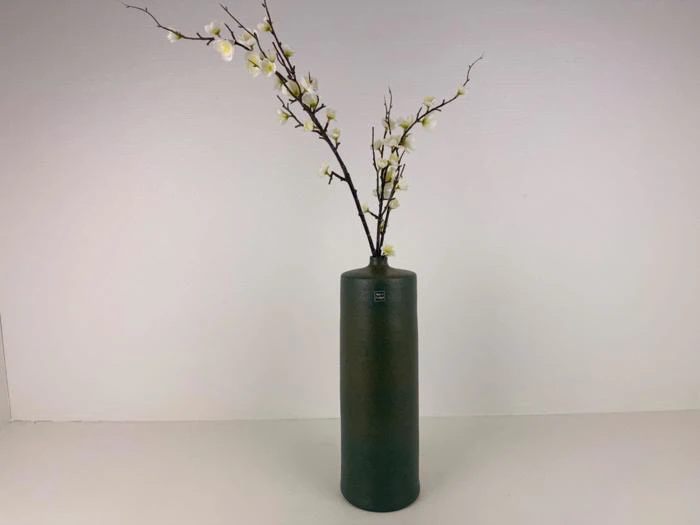 En høj, mørkegrøn keramikvase holder to lange grene med små hvide blomster mod en almindelig hvid baggrund.
