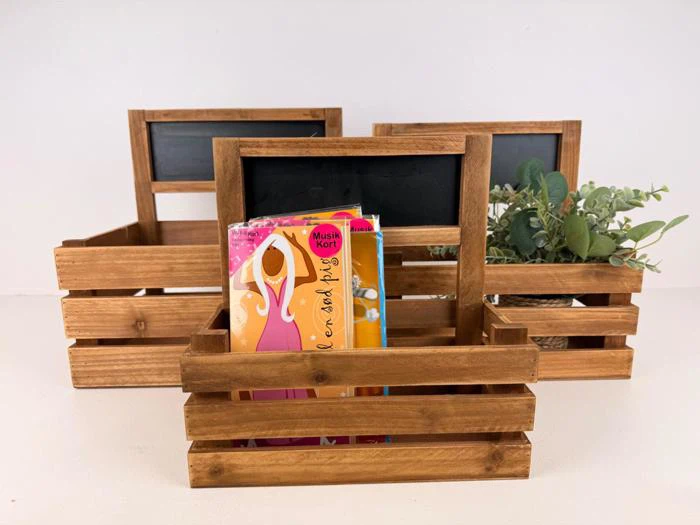 Tre trækasser med kridtetiketter; en indeholder farverige kort, en anden har en potteplante, og den tredje er tom. Alle er placeret på en hvid overflade mod en almindelig baggrund.