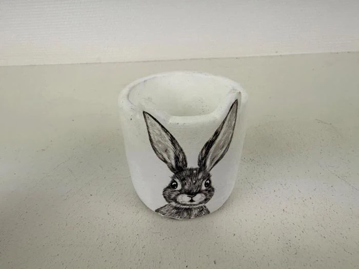 En hvid keramiklysestage med en sort illustration af et kaninansigt og store ører på siden, placeret på en lys overflade.