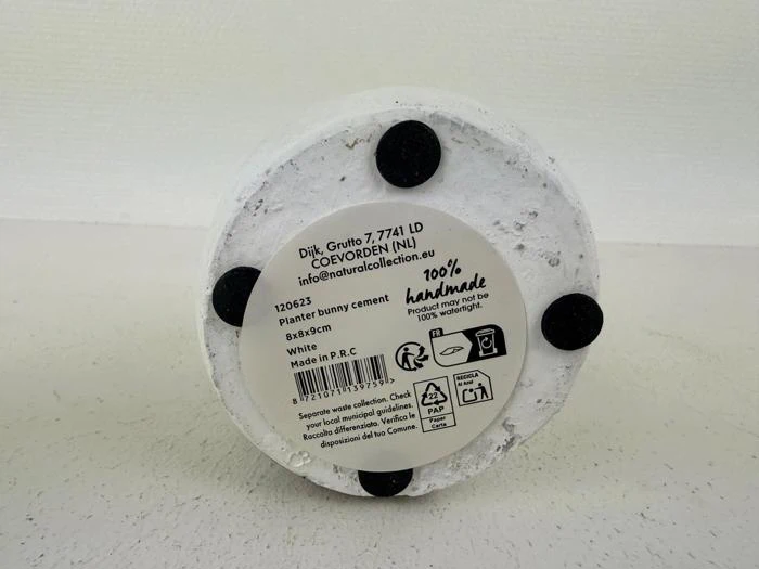 Bunden af en hvid cementpotte med fire sorte puder og en etiket med produktoplysninger, herunder 100 % håndlavet, Planter bunny cement og produktionsoplysninger.