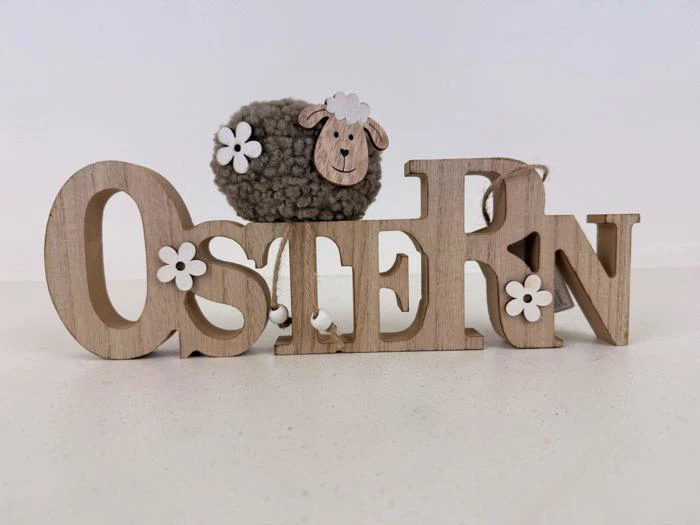 Træbogstaver, der staver "OSTERN" med hvide blomsterdekorationer, og et lille plysfår med et smilende ansigt bag bogstaverne.