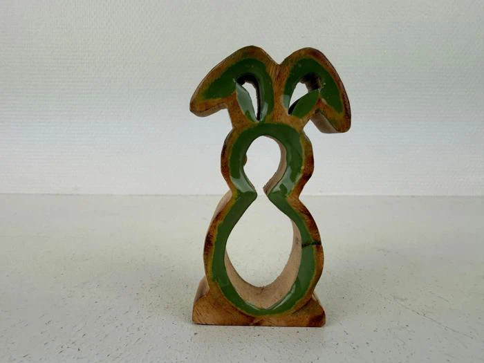 En silhuet af en påskehare i træ med grønmalede kanter og udskæringer står oprejst på en hvid overflade mod en struktureret hvid baggrund.